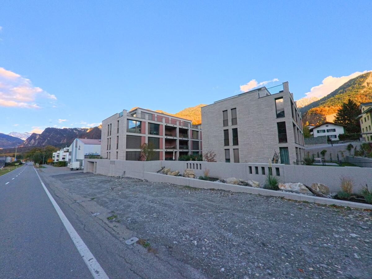 Entlang einer Hauptstrasse in Trimmis (7203, Kanton Graubünden) stehen moderne Mehrfamilienhäuser mit klaren Fassaden, vor denen sich der Blick zu den herbstlich beleuchteten Berghängen öffnet.