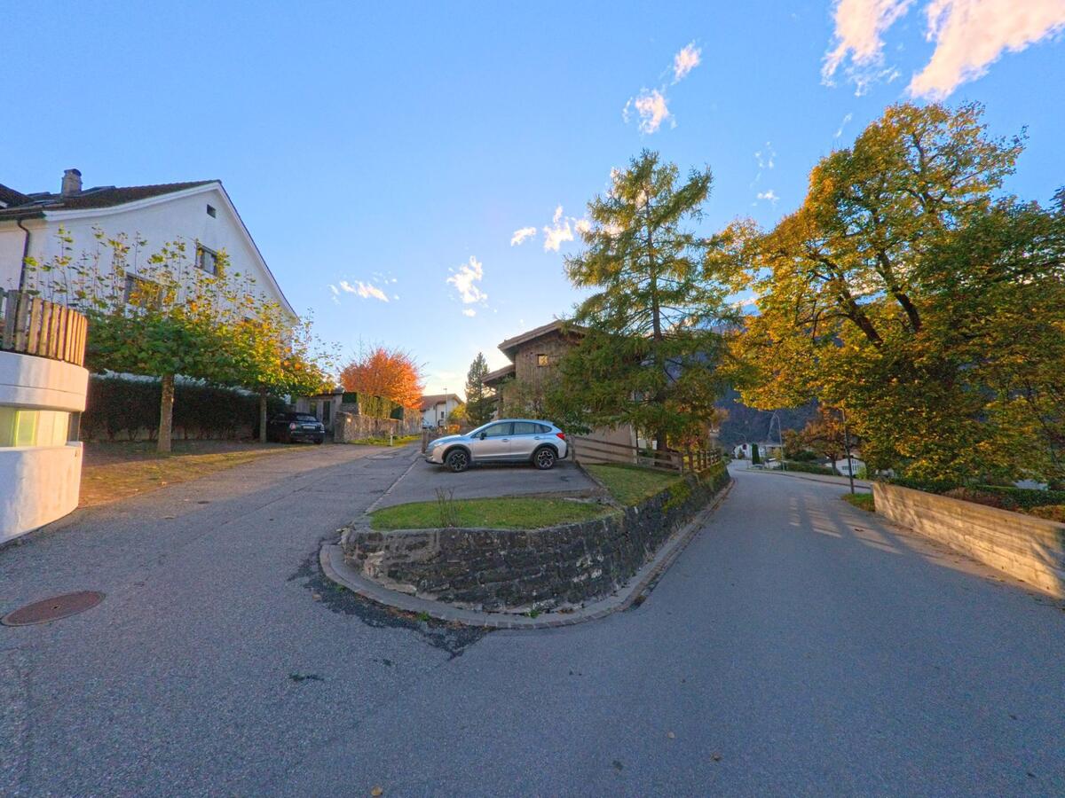 Zwischen Wohnhäusern in Trimmis (7203, Kanton Graubünden) verzweigt sich eine ruhige Quartierstrasse, begleitet von herbstlich gefärbten Bäumen und kleinen Gärten.