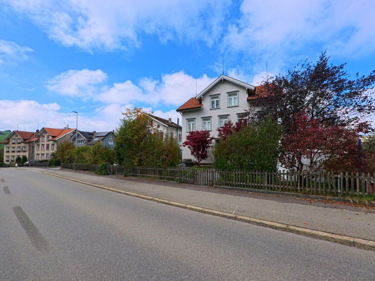 Entlang einer ruhigen Strasse in Trogen (9043, Kanton Appenzell Ausserrhoden) stehen gepflegte Wohnhaeuser und herbstlich gefaerbte Straeucher.