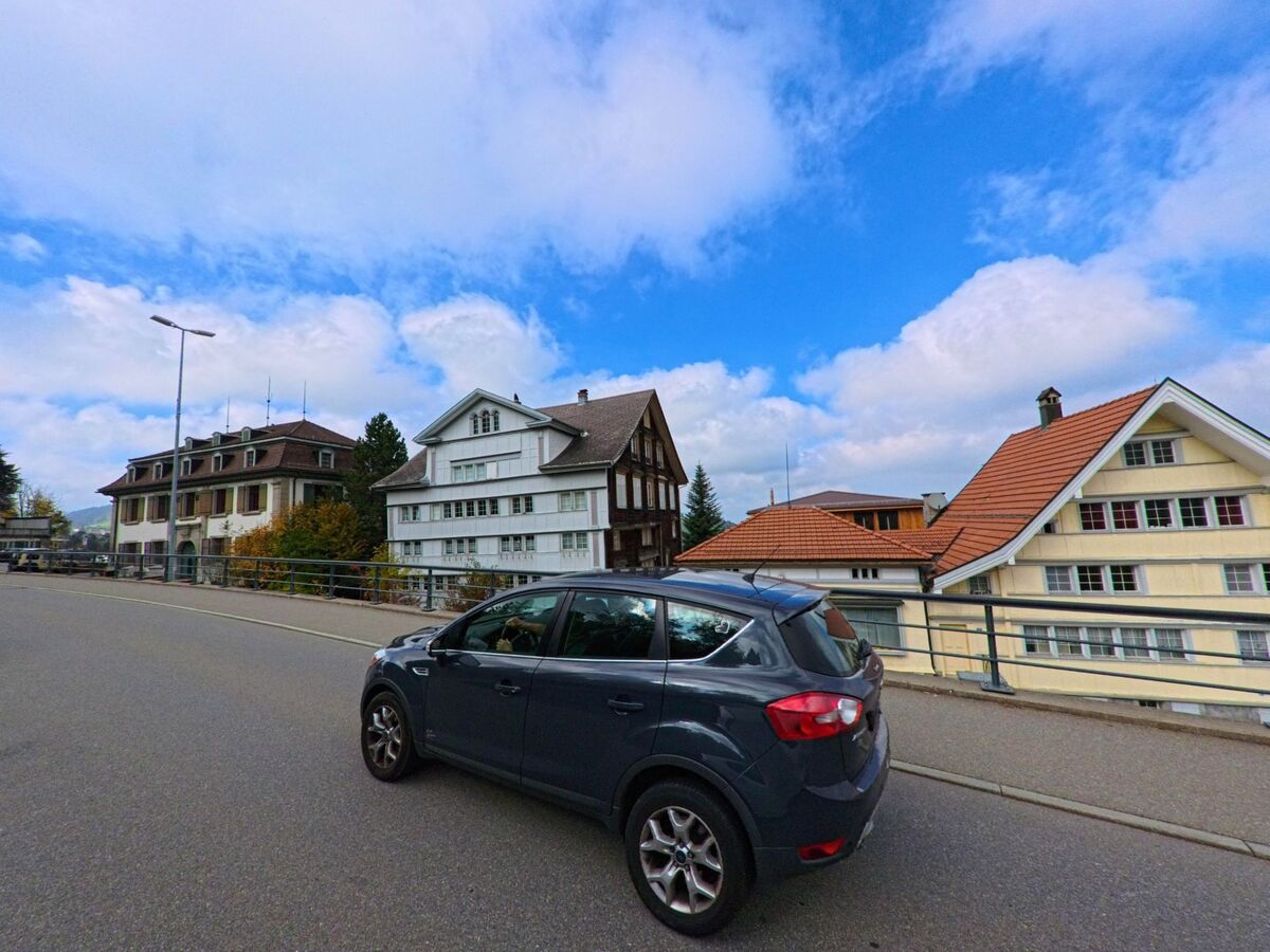 An einer leicht ansteigenden Strasse in Trogen (9043, Kanton Appenzell Ausserrhoden) reihen sich traditionelle Haeuser mit hellen Fassaden unter einem blauen Himmel.