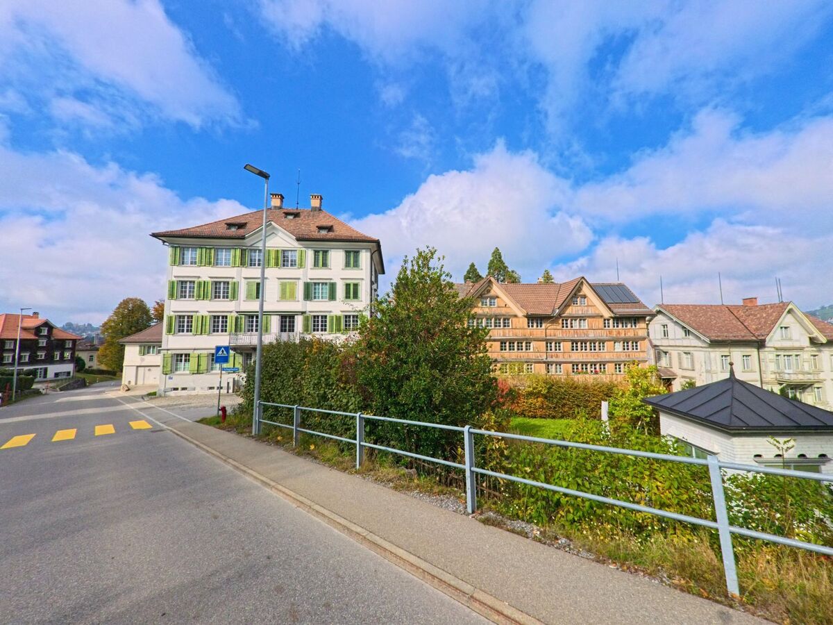An einer stillen Strasse in Trogen (9043, Kanton Appenzell Ausserrhoden) stehen helle Haeuser mit gruenen und holzfarbenen Fassaden vor blauem Himmel.
