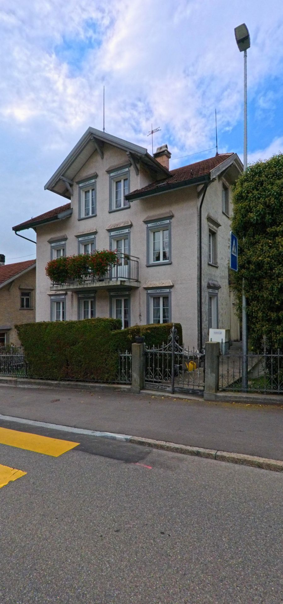 Vor einem hellen Wohnhaus in Trogen (9043, Kanton Appenzell Ausserrhoden) stehen eine Hecke und ein schmiedeeiserner Zaun entlang der Strasse.