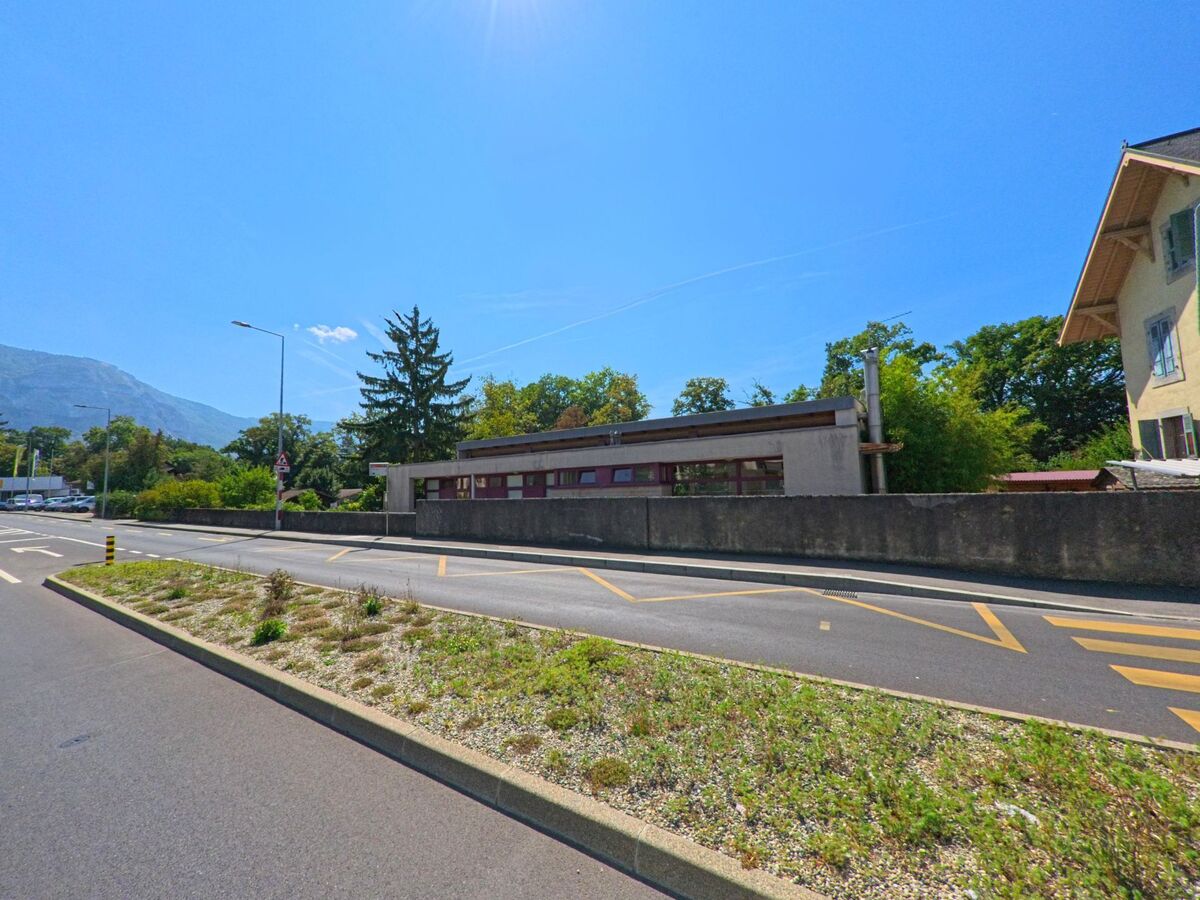 Eine Hauptstrasse in Troinex (1256, Kanton Genf) verlaeuft an einem flachen Gebaeude vorbei, im Hintergrund sind Baeume und ein Berghang sichtbar.