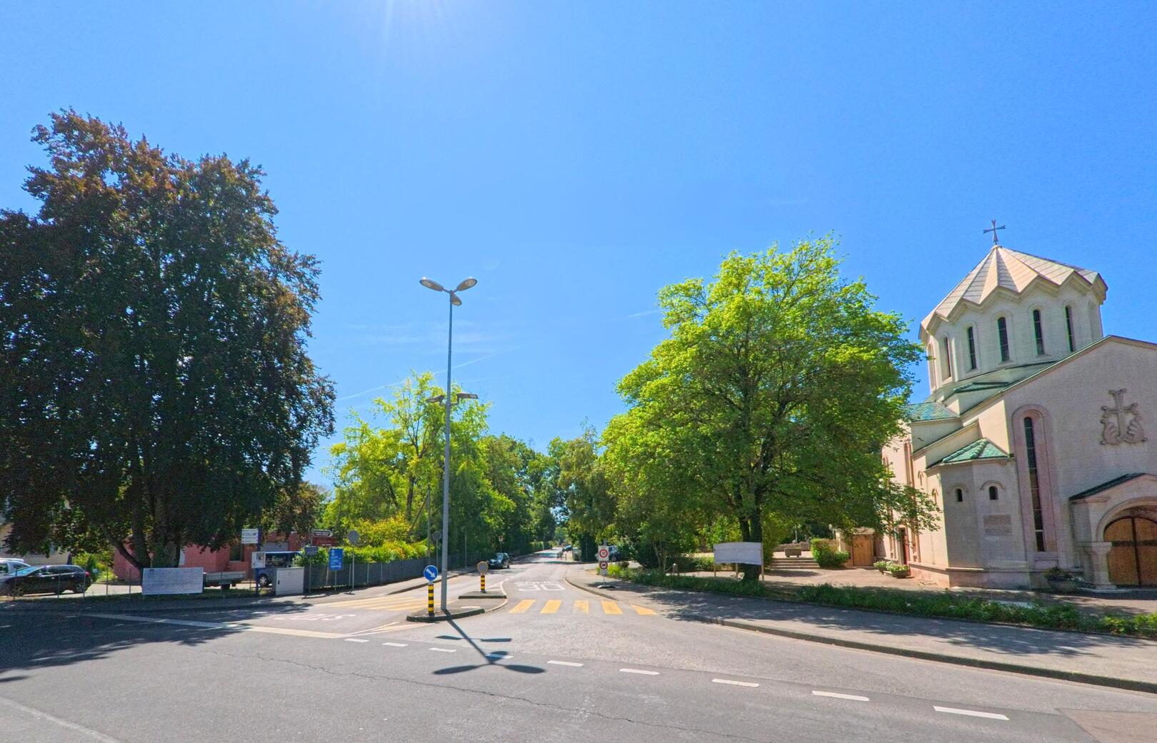 An einer Strassenkreuzung in Troinex (1256, Kanton Genf) steht eine Kirche mit markantem Turm neben grossen Baeumen.