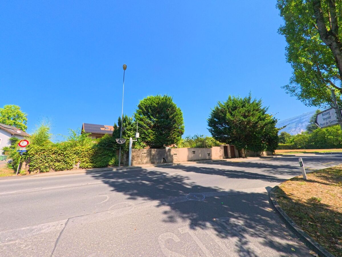 An einer Kreuzung in Troinex (1256, Kanton Genf) fuehrt eine Strasse zwischen Mauern und dichten Baeumen vorbei, im Hintergrund ist ein Huegel sichtbar.