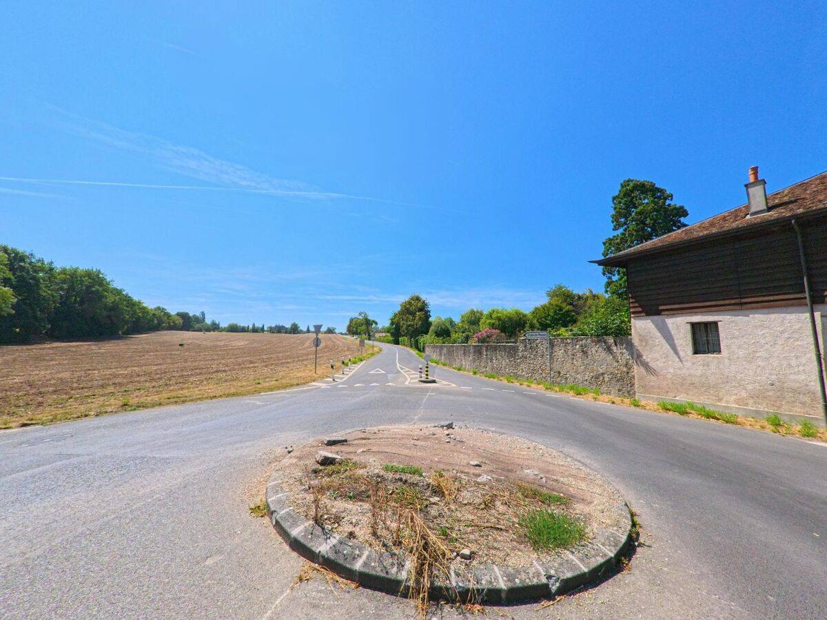 Eine schmale Landstrasse fuehrt in Troinex (1256, Kanton Genf) zwischen Feldern und einer Steinmauer an einem Bauernhaus vorbei.