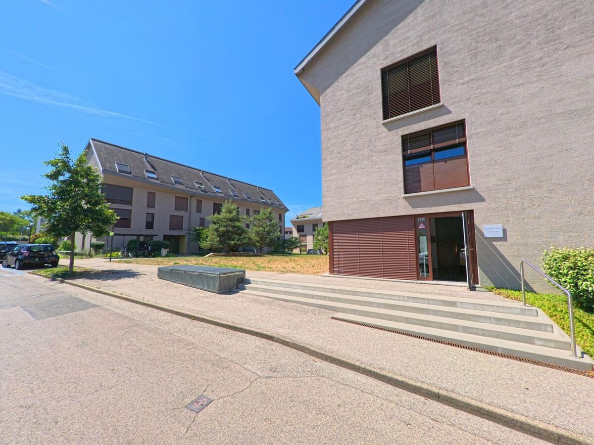 Ein modernes Verwaltungsgebaeude in Troinex (1256, Kanton Genf) liegt an einer Strasse mit weiteren Wohnbauten im Hintergrund.