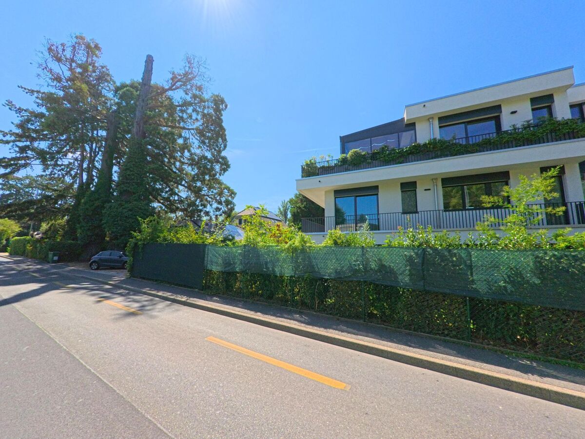 In Troinex (1256, Kanton Genf) steht ein modernes Wohnhaus mit grossen Balkonen neben einer Baumreihe an der Strasse.