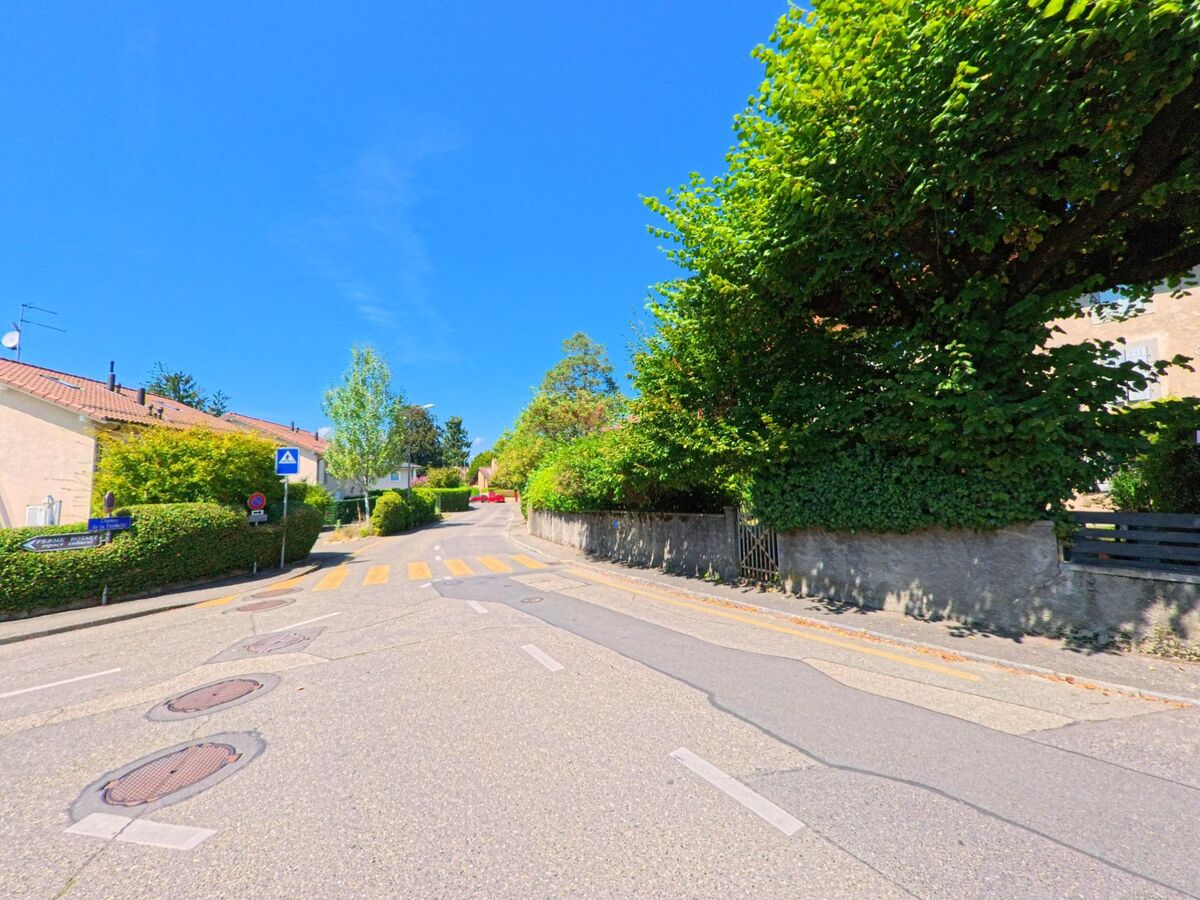 Eine Wohnstrasse in Troinex (1256, Kanton Genf) fuehrt vorbei an Haeusern mit Hecken und einem markierten Fussgaengerstreifen.