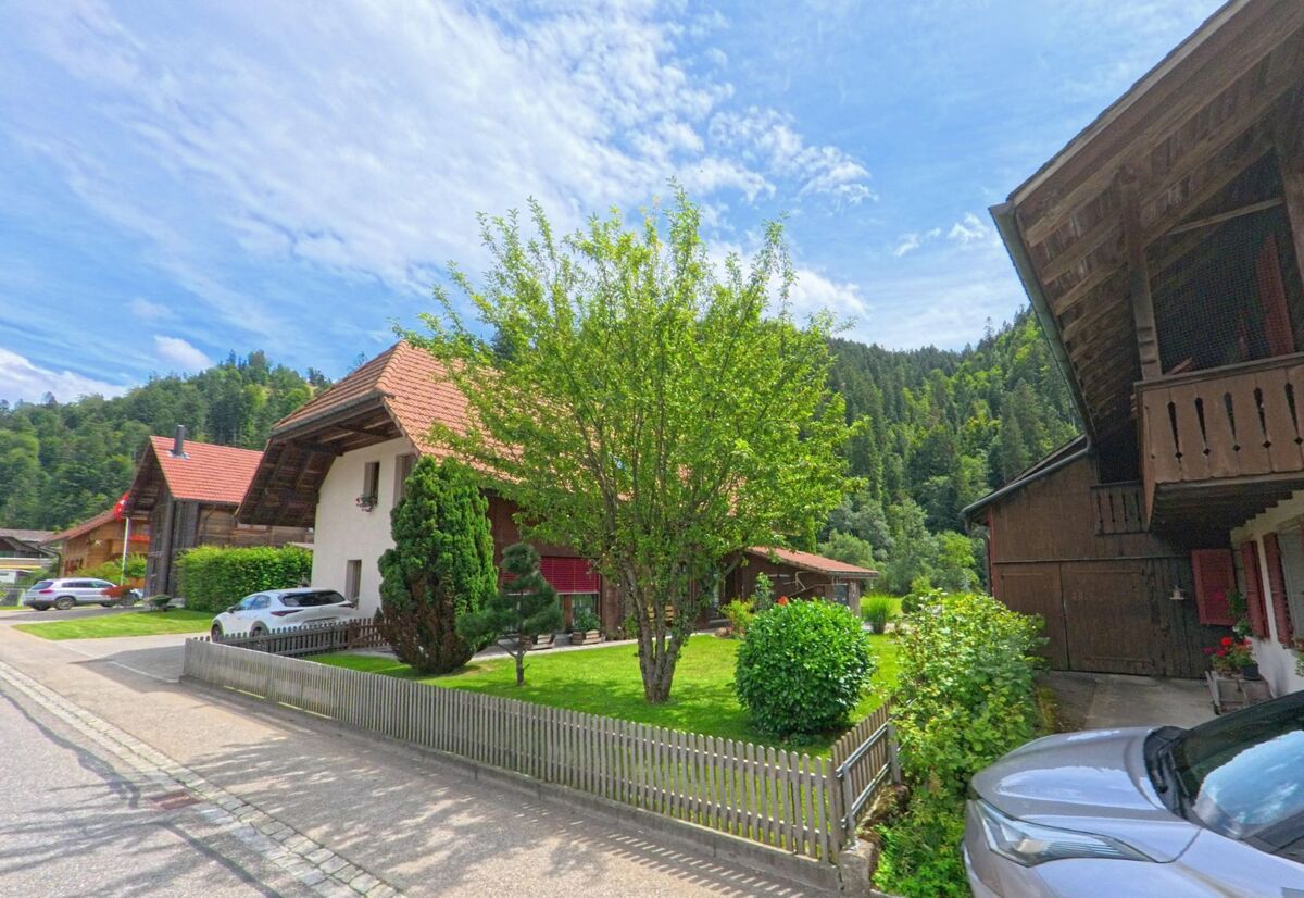 In Trub (3556, Kanton Bern) säumen gepflegte Einfamilienhäuser mit Gärten und Holzzäunen eine ruhige Strasse am Waldrand, eingebettet in eine grüne Hügellandschaft.