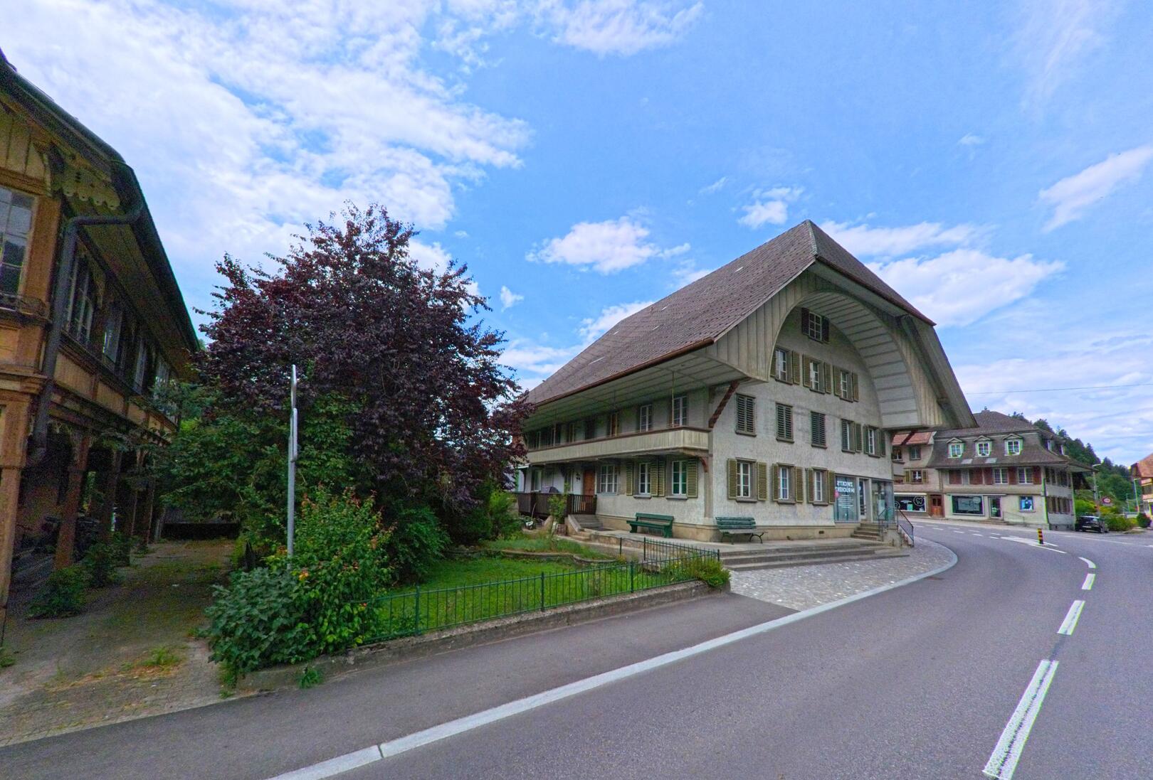 Ein traditionelles Bauernhaus mit grossem Walmdach prägt das Ortsbild von Trubschachen (3555, Kanton Bern) an einer sanft geschwungenen Strasse.
