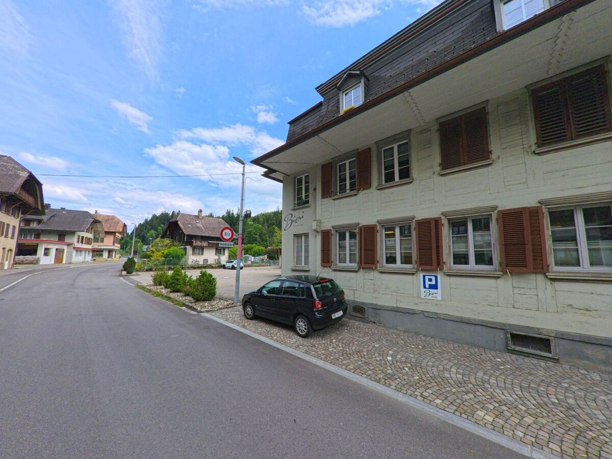 Das Bild zeigt ein charakteristisches Emmentaler Haus mit breitem Walmdach im Ortskern von Trubschachen (3555, Kanton Bern), eingerahmt von weiteren traditionellen Gebäuden entlang der Hauptstrasse.