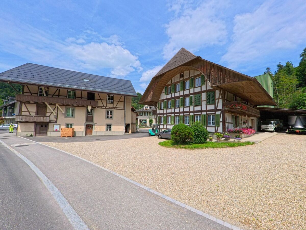 Zwei traditionelle Gebäude mit Fachwerkfassade und grosszügigem Kiesvorplatz stehen in Trubschachen (3555, Kanton Bern) am Rand eines bewaldeten Hangs, begleitet von landwirtschaftlichen Fahrzeugen.
