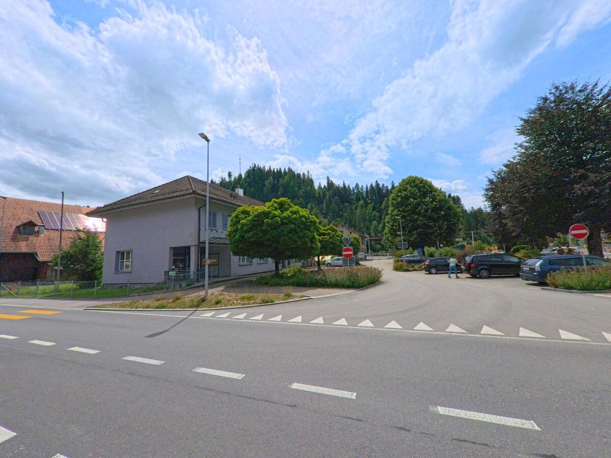 Ein kleiner Platz mit Parkplätzen und Einbahnstrassenregelung liegt in Trubschachen (3555, Kanton Bern) vor einem Wohnhaus und wird von mehreren Bäumen und Verkehrszeichen gesäumt.