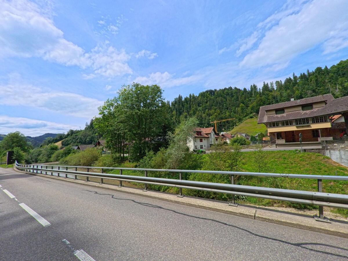 Am Ortsrand von Trubschachen (3555, Kanton Bern) verläuft eine Strasse mit Leitplanke entlang eines grünen Hangs, dahinter liegen verstreute Häuser und bewaldete Hügel.