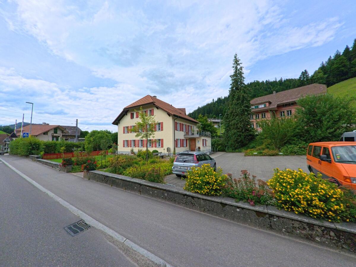 Ein helles Wohnhaus mit roten Fensterläden steht in Trubschachen (3555, Kanton Bern) an einer ruhigen Strasse, umgeben von gepflegten Gärten und einem kleinen Parkplatz.