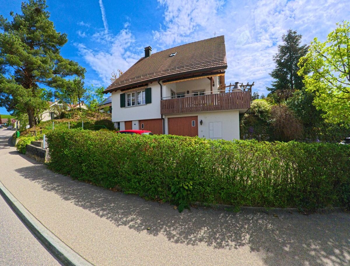 Ein Einfamilienhaus mit grossem Holzbalkon und gruener Hecke steht in Truellikon (8466, Kanton Zuerich) an einer ruhigen Wohnstrasse.