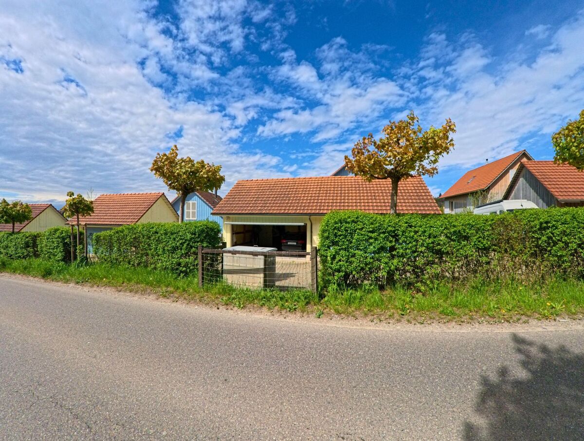 Hinter einer dichten Hecke in Truellikon (8466, Kanton Zuerich) stehen kleine Gebaeude mit roten Ziegeldachern und gepflegten Baeumen.