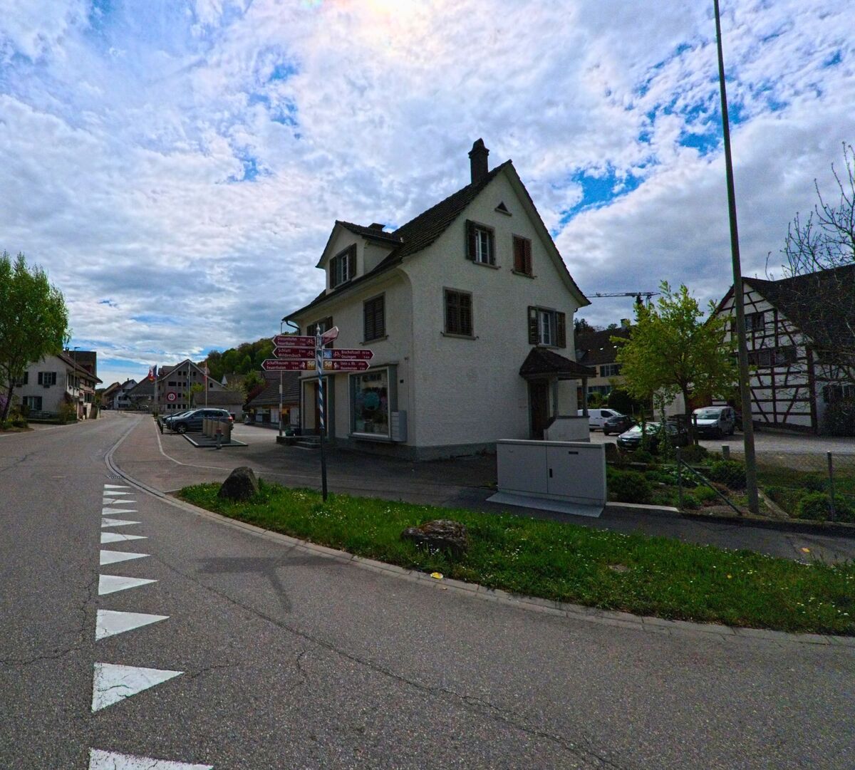 An einer Kreuzung in Truellikon (8466, Kanton Zuerich) steht ein weisses Wohn- und Geschaeftshaus neben Wegweisern fuer verschiedene Ziele.