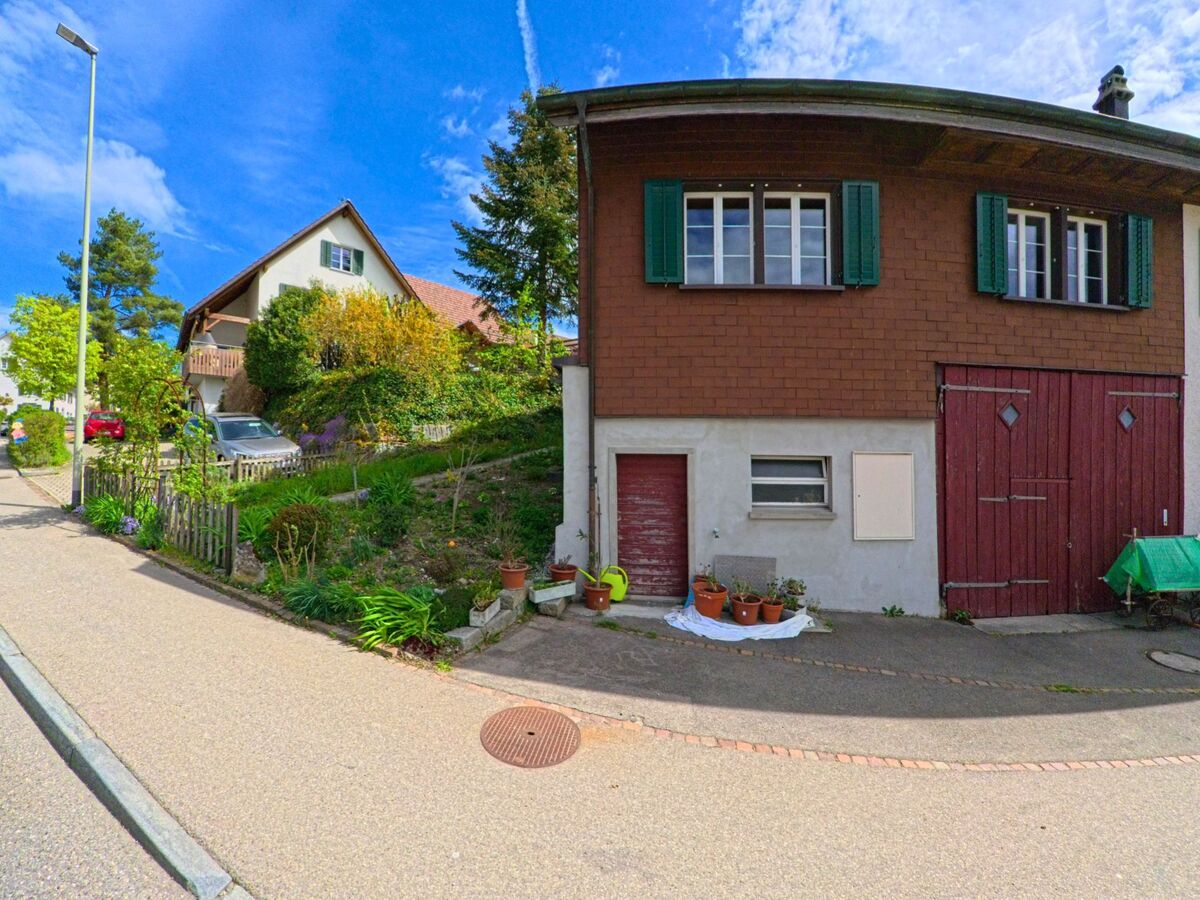 Ein Wohnhaus mit rotem Scheunentor und gruene Fensterlaeden steht in Truellikon (8466, Kanton Zuerich) an einer ruhigen Strasse mit angrenzendem Garten.