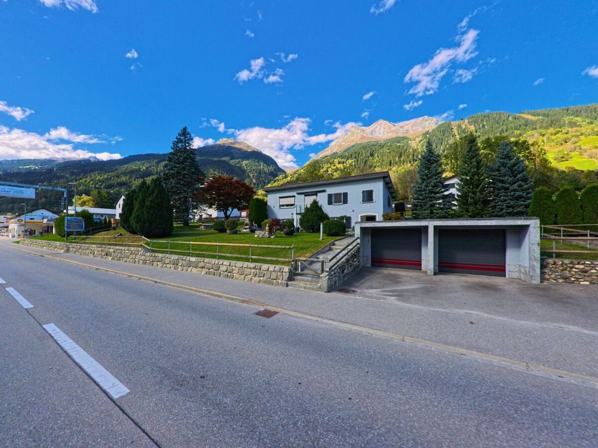 Im Zentrum von Trun (7166, Kanton Graubuenden) stehen zwei auffaellige Gebaeude mit gruenen und schwarz-weissen Fensterlaeden entlang einer gepflasterten Strasse.