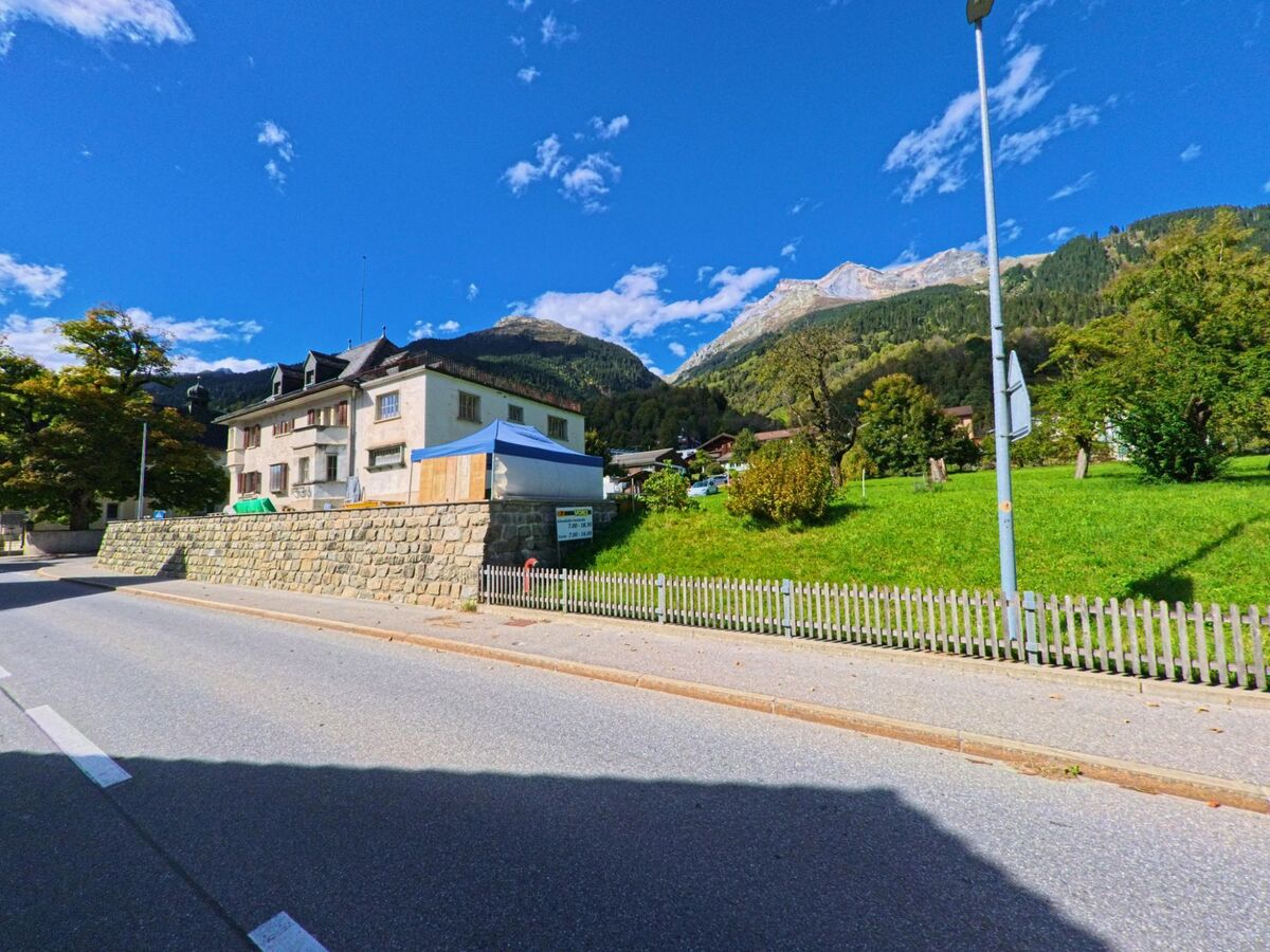 Ein helles Gebaeude mit Mansarddach steht in Trun (7166, Kanton Graubuenden) an einer ruhigen Strasse, umgeben von gruenen Wiesen und Bergen im Hintergrund.