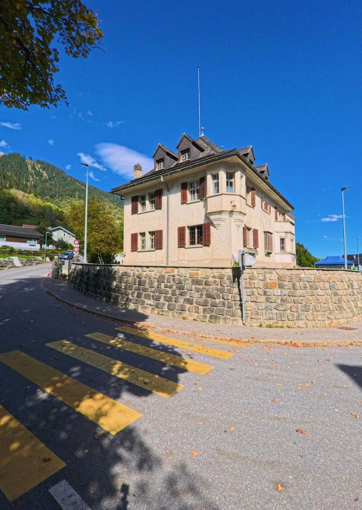 Ein historisches Gebaeude mit Mansarddach und roten Fensterlaeden steht in Trun (7166, Kanton Graubuenden) an einer Strassenecke mit Fussgaengerstreifen.