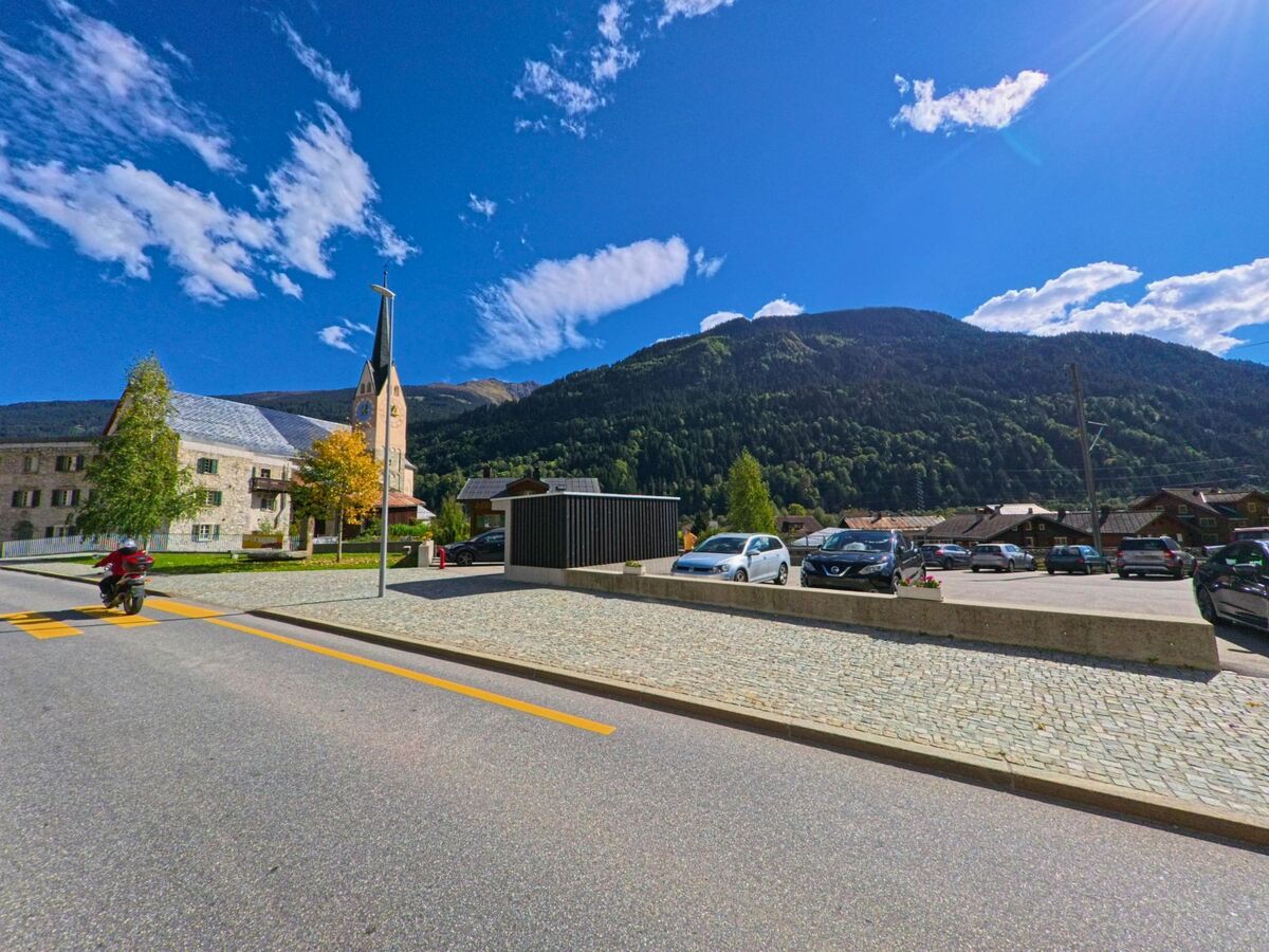 Vor der Bergkulisse von Trun (7166, Kanton Graubuenden) steht eine Kirche mit spitzem Turm neben einem Parkplatz und mehreren Wohnhaeusern bei klarem Himmel.