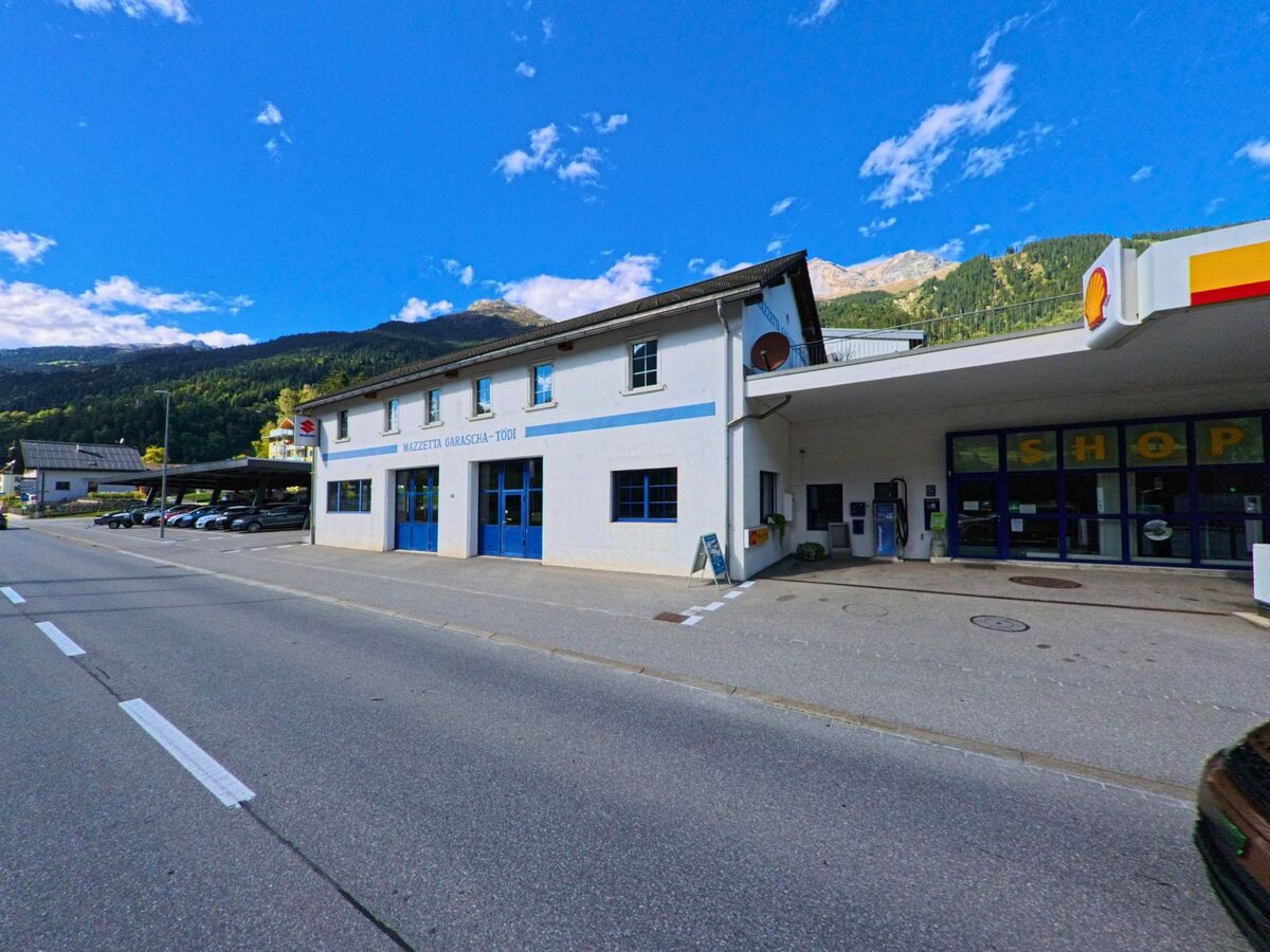 Im Zentrum von Trun (7166, Kanton Graubuenden) stehen zwei auffaellige Gebaeude mit gruenen und schwarz-weissen Fensterlaeden entlang einer gepflasterten Strasse.