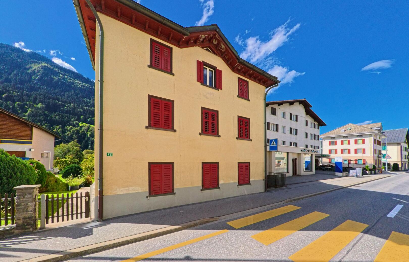 An der Hauptstrasse von Trun (7166, Kanton Graubuenden) steht ein gelbes Wohnhaus mit roten Fensterlaeden neben weiteren Gebaeuden vor der Kulisse bewaldeter Berghaenge.