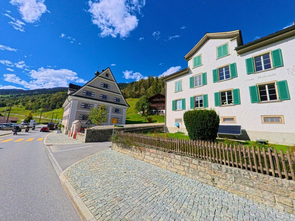 Im Zentrum von Trun (7166, Kanton Graubuenden) stehen zwei auffaellige Gebaeude mit gruenen und schwarz-weissen Fensterlaeden entlang einer gepflasterten Strasse.