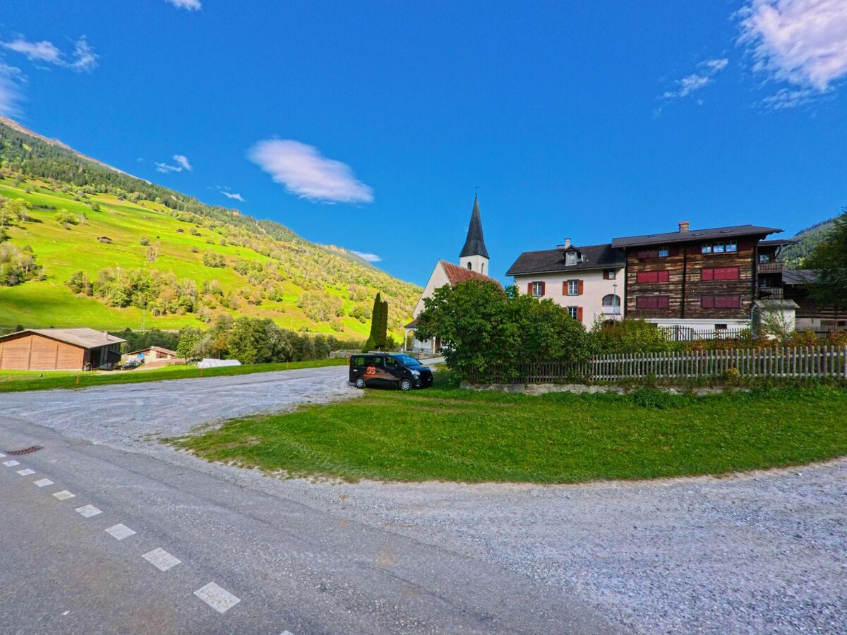 Im Zentrum von Trun (7167, Kanton Graubuenden) steht eine Kirche mit spitzem Turm, umgeben von traditionellen Wohnhaeusern und einer grünen Berglandschaft im Hintergrund.