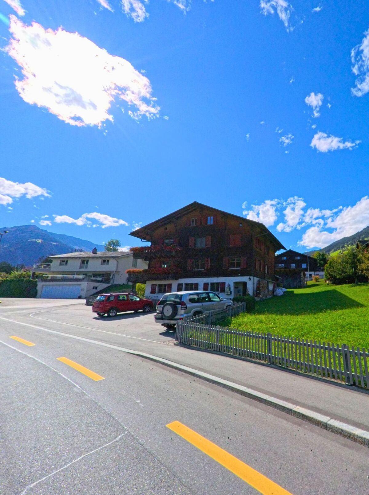 Am Dorfrand von Trun (7172, Kanton Graubuenden) stehen mehrere gepflegte Wohnhaeuser mit Holzbalkonen und Fensterlaeden vor einer gruenen Hanglandschaft.