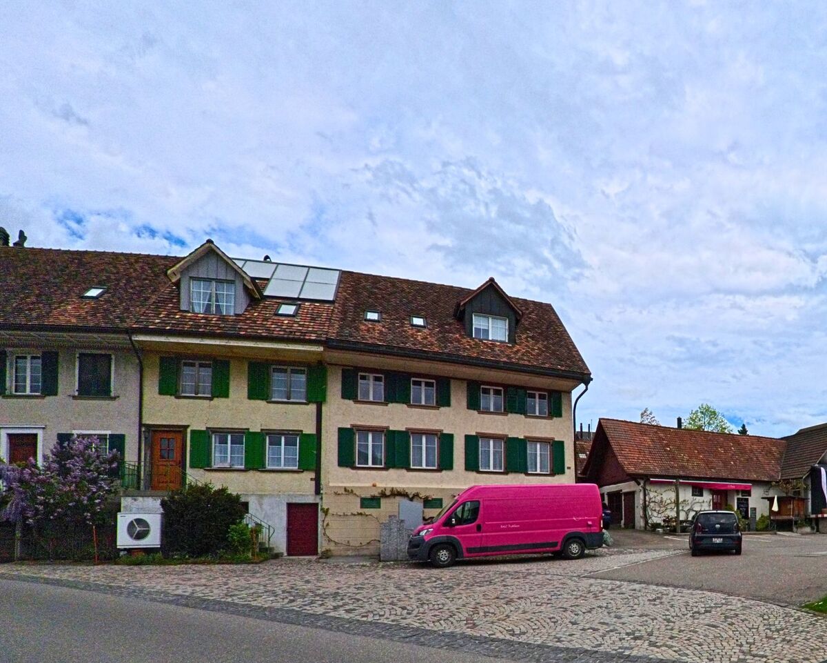In Truttikon (8467, Kanton Zürich) steht ein langgezogenes Wohnhaus mit grün gestrichenen Fensterläden und Solarpanels auf dem Dach an einem gepflasterten Platz.