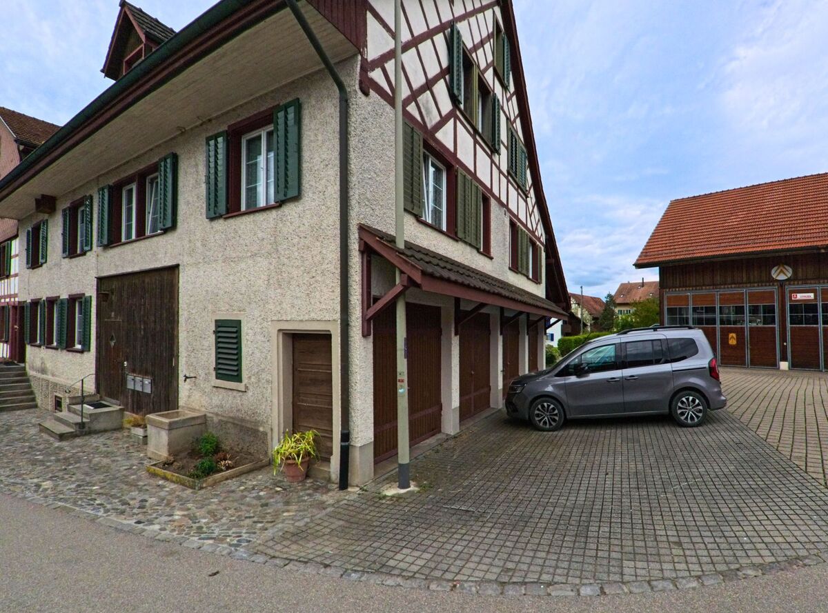  In Truttikon (8467, Kanton Zürich) steht ein traditionelles Wohnhaus mit grünen Fensterläden neben dem Gebäude der Feuerwehr mit grossen Toren.