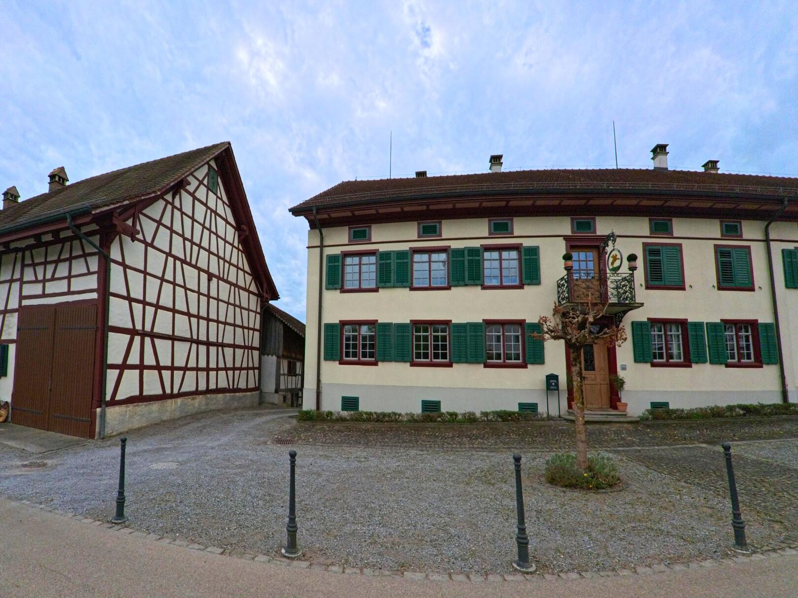 Vor dem Gemeindehaus in Truttikon (8467, Kanton Zürich) mit grün gestrichenen Fensterläden und Fachwerkgebäude daneben liegt ein gepflasterter Platz mit junger Baumbepflanzung.