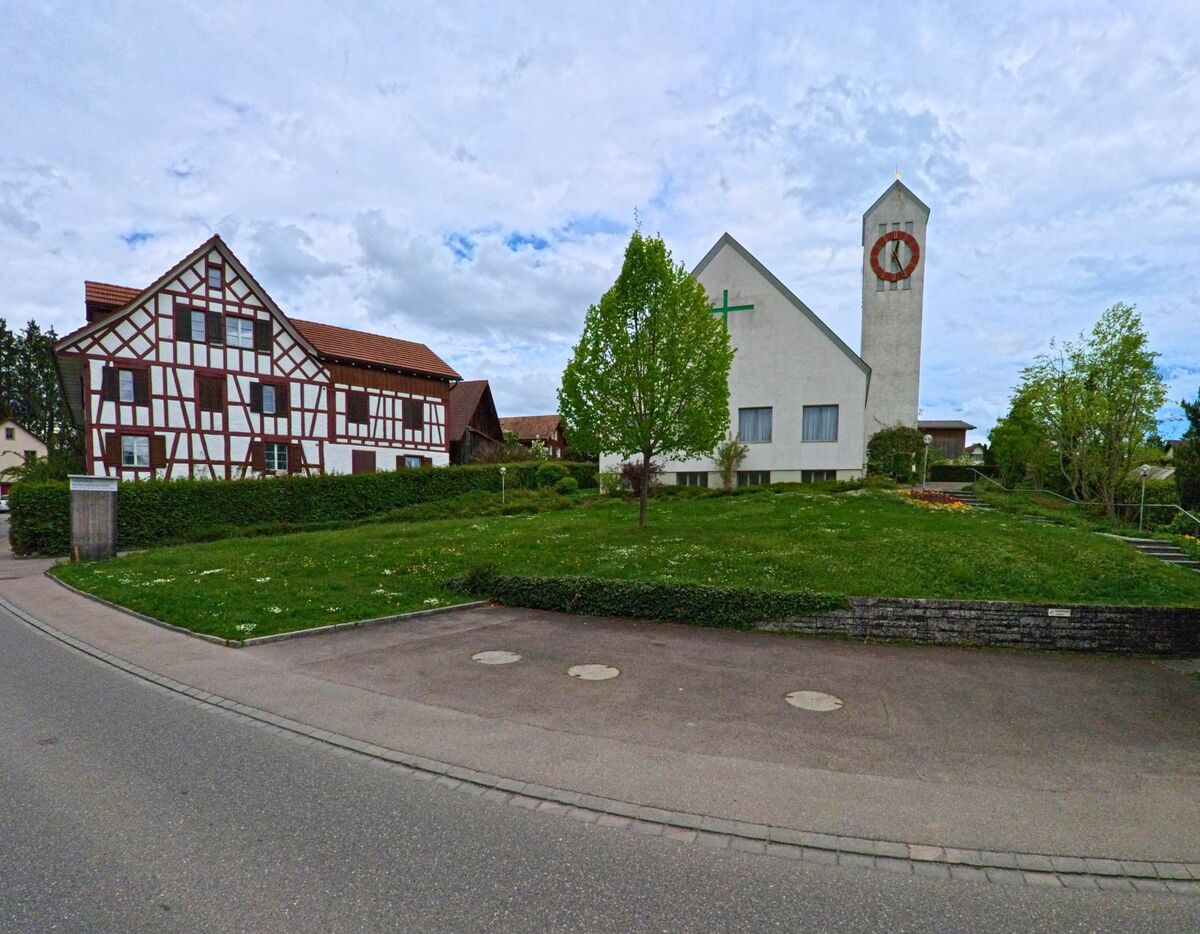 In Truttikon (8467, Kanton Zürich) stehen eine moderne Kirche mit markantem Glockenturm und ein traditionelles Fachwerkhaus nebeneinander an einer ruhigen Strasse.