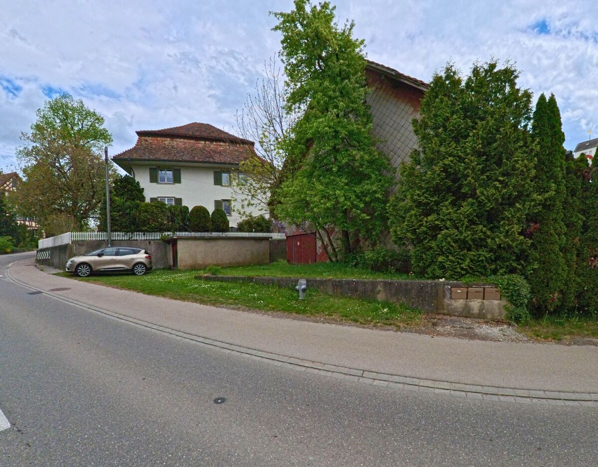An einer sanft geschwungenen Strasse in Truttikon (8467, Kanton Zürich) steht ein markantes Wohnhaus mit Walmdach, umgeben von gepflegtem Grün und alten Bäumen.