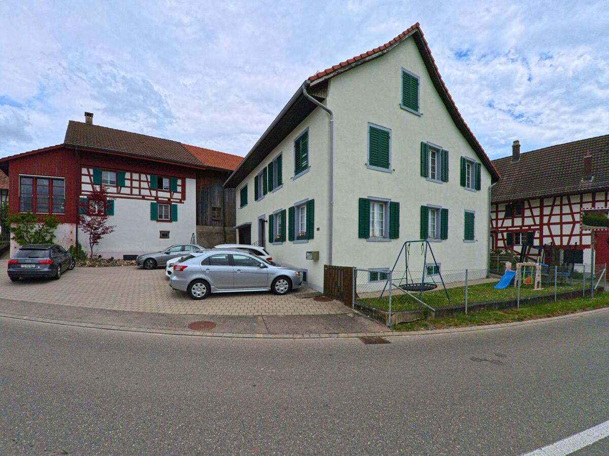 In Truttikon (8467, Kanton Zürich) säumen Wohnhäuser mit grünen Fensterläden und sichtbarem Fachwerk eine leicht geschwungene Strasse, daneben ein kleiner umzäunter Spielbereich.