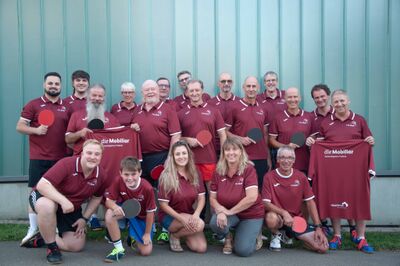 In Muri (5630, Kanton Aargau) sitzt und steht der TTC Muri AG als grosse Gruppe in einheitlichen weinroten Trikots mit Tischtennisschlaegern vor einer modernen Metallfassade.