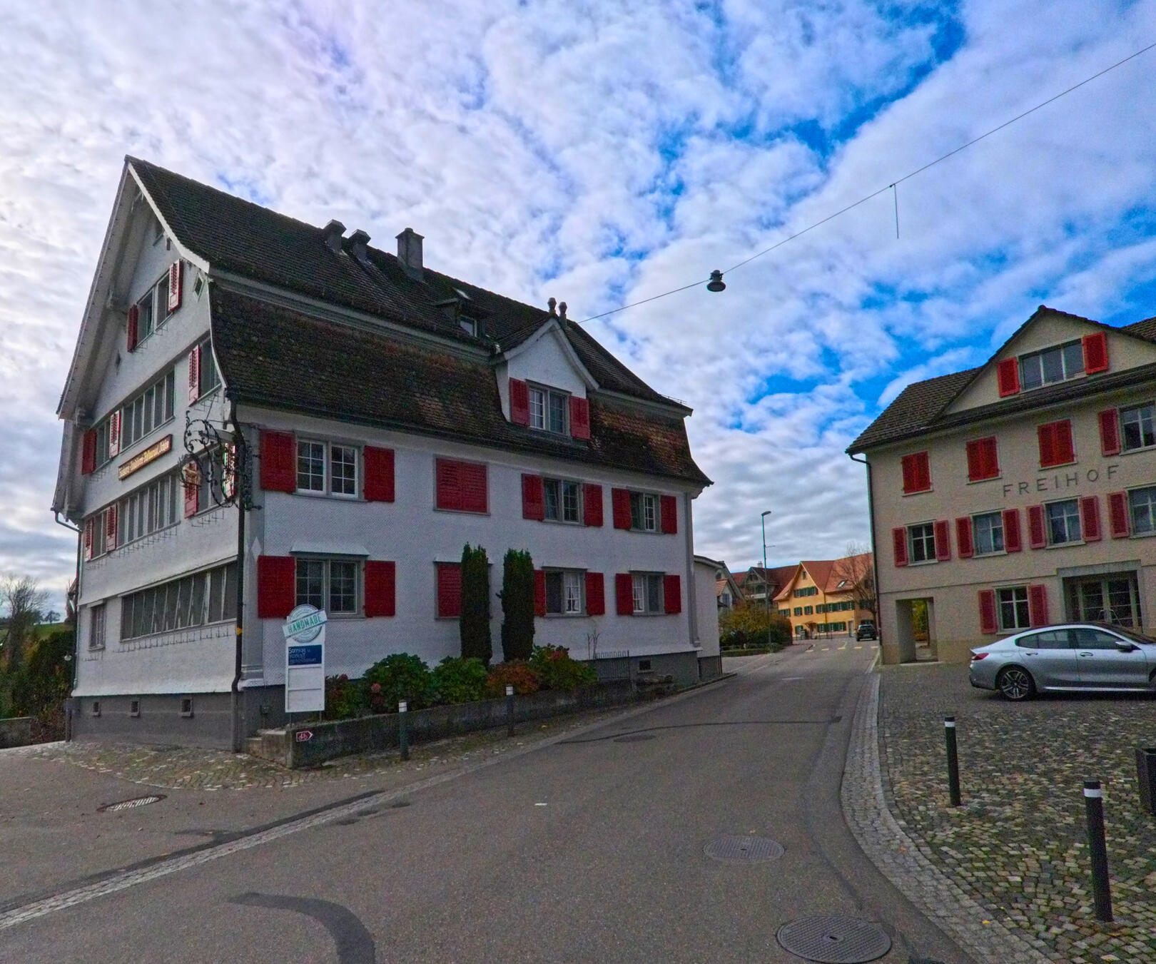 An einer ruhigen Strasse in Tuebach (9402, Kanton St. Gallen) stehen traditionelle Wohnhaeuser mit roten Fensterlaeden unter einem wolkigen Himmel.