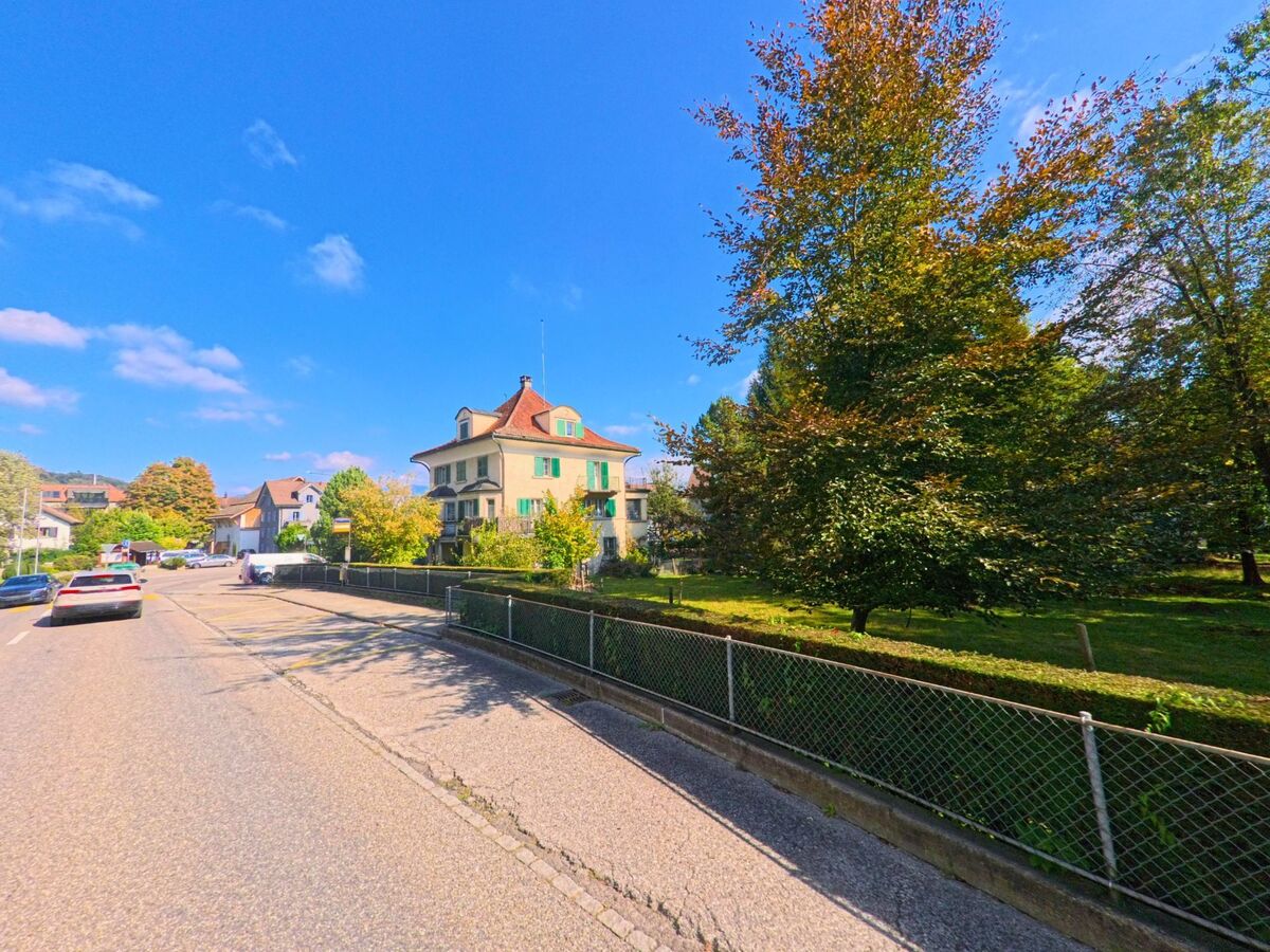 In Tuggen (8856, Kanton Schwyz) verläuft eine ruhige Strasse an einem grossen Garten mit Baumbestand vorbei, waehrend ein markantes Haus mit gruenen Laeden im Sonnenlicht steht.