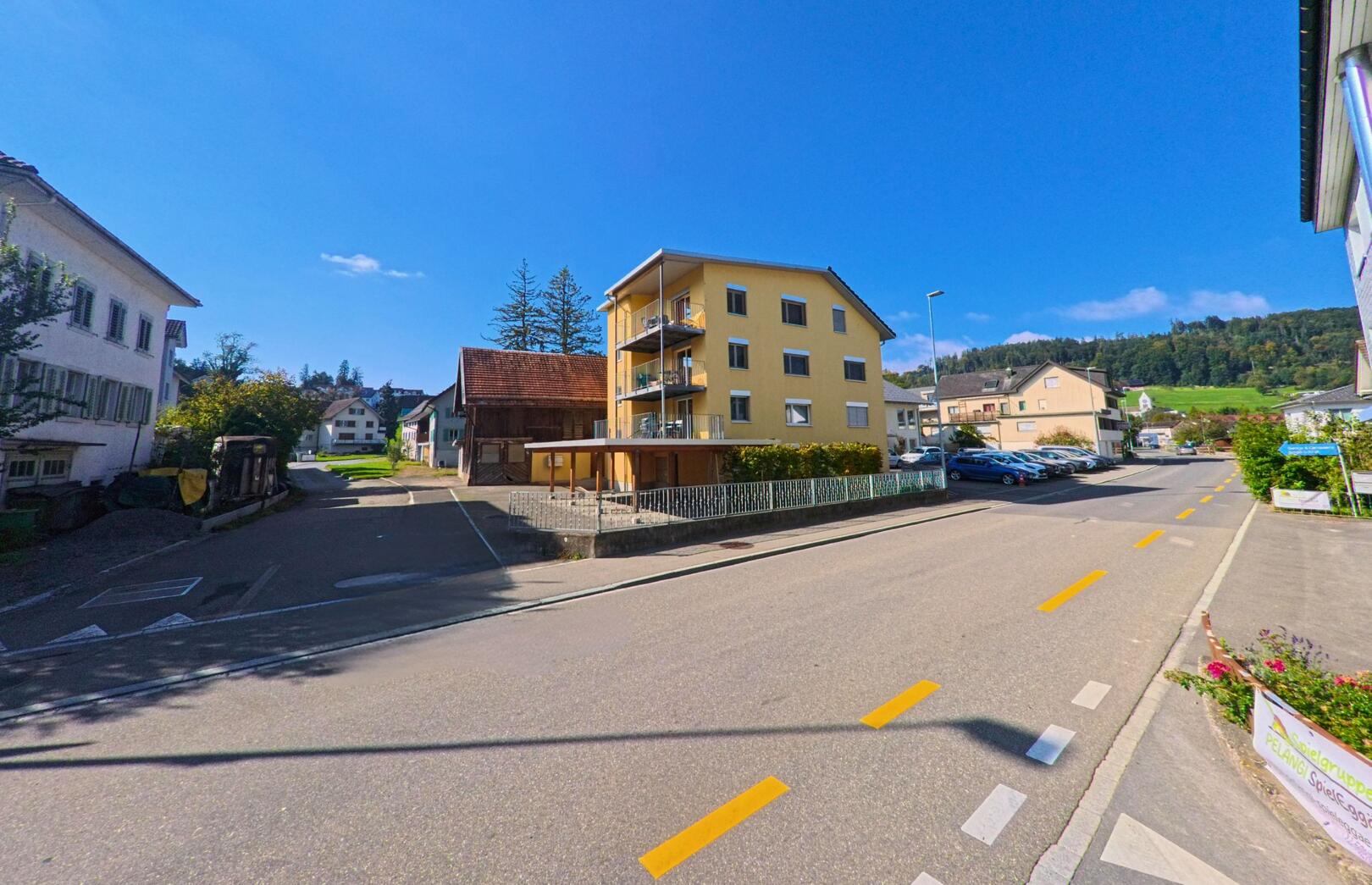In einer sonnigen Strassenszene in Tuggen (8856, Kanton Schwyz) steht ein gelbes Wohnhaus neben alten und neuen Gebaeuden vor dem bewaldeten Hang.