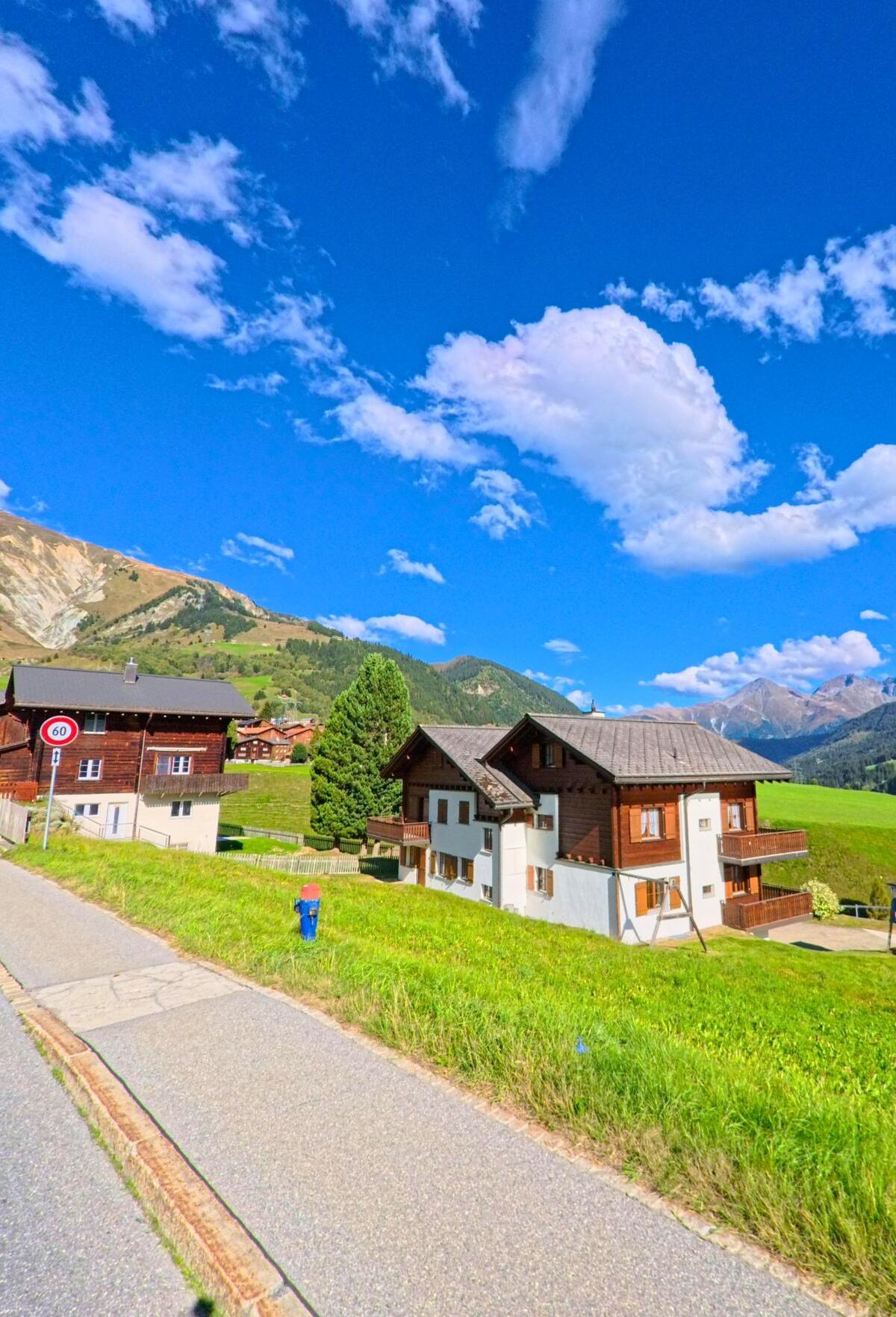 In Tujetsch (7187, Kanton Graubuenden) stehen traditionelle Holzhäuser mit hellen Fassaden vor einer Bergkulisse unter blauem Himmel.