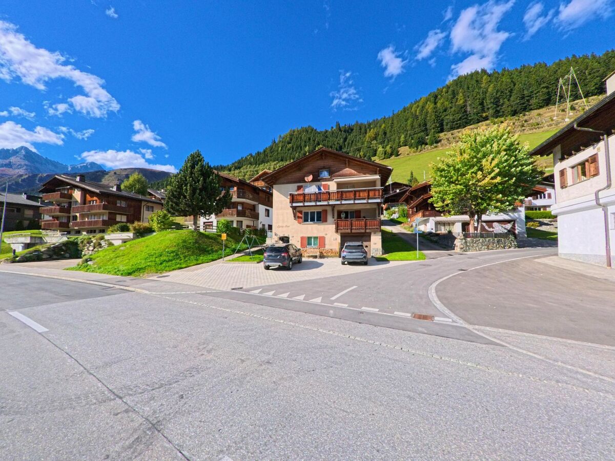 In Tujetsch (7188, Kanton Graubuenden) stehen mehrere Chalets mit Holzbalkonen und gepflegten Gaerten an einer leicht ansteigenden Strasse vor bewaldeten Haengen.