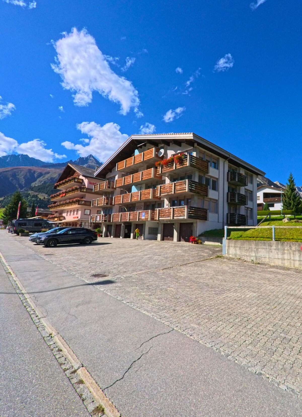 Ein mehrstöckiges Wohnhaus mit Holzbalkonen steht in Tujetsch (7188, Kanton Graubuenden) an einer gepflasterten Strasse mit Blick auf die Berge.