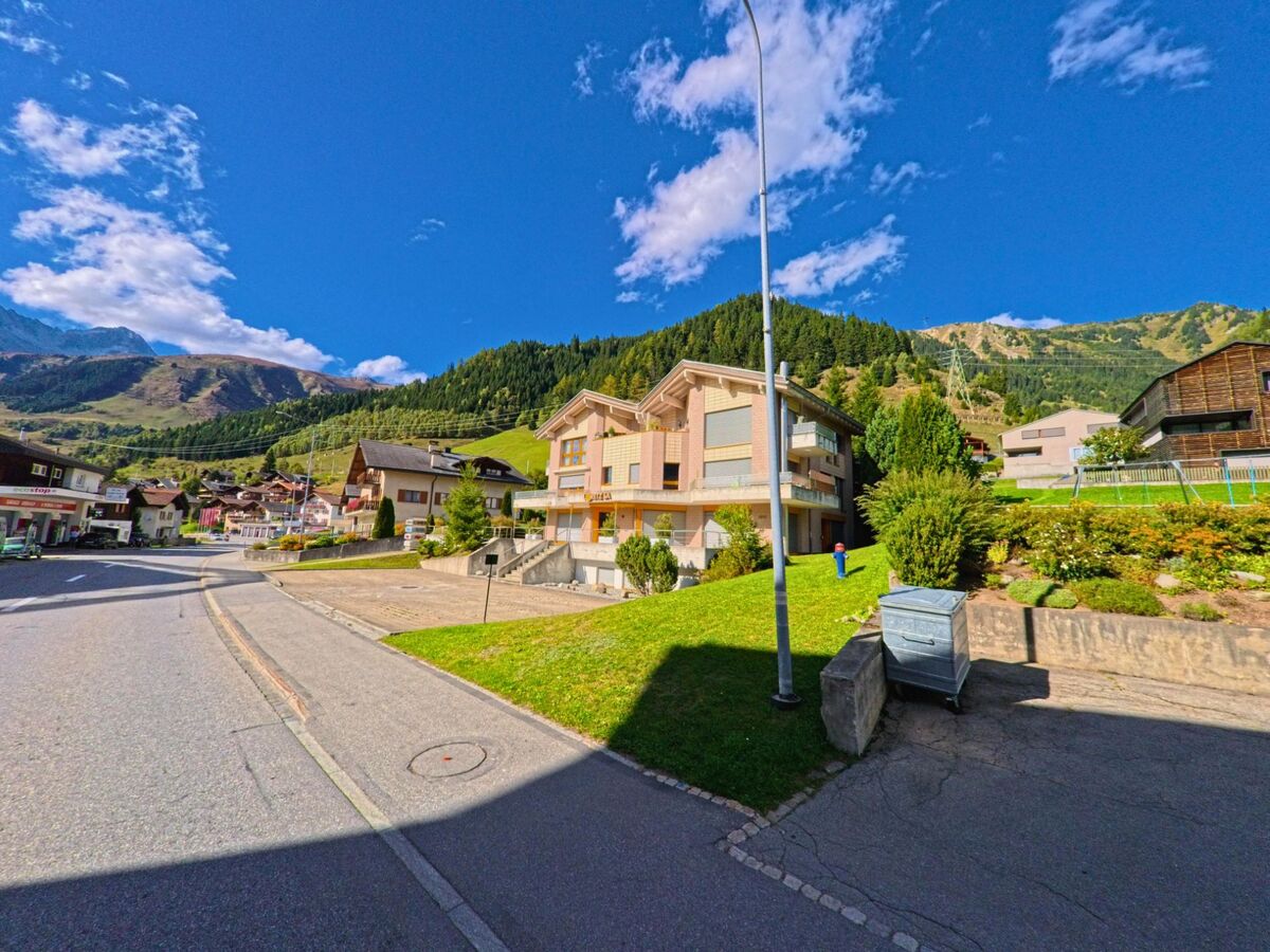 In Tujetsch (7188, Kanton Graubuenden) reihen sich moderne Wohnhaeuser und traditionelle Bauten entlang einer Strasse am Hang mit Blick auf bewaldete Berge.