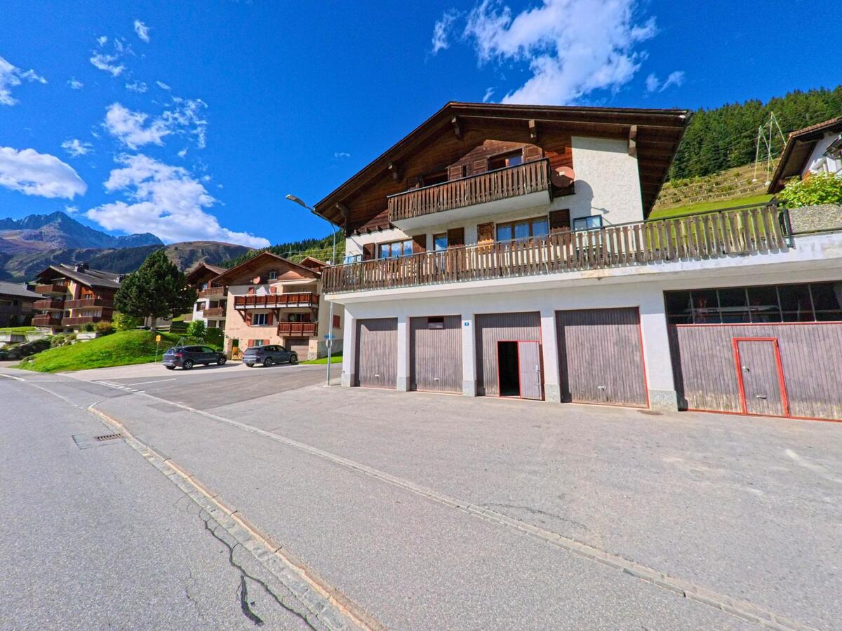 Mehrere Wohnhaeuser mit Holzbalkonen liegen in Tujetsch (7188, Kanton Graubuenden) an einer ruhigen Strasse mit Blick auf die Berglandschaft.