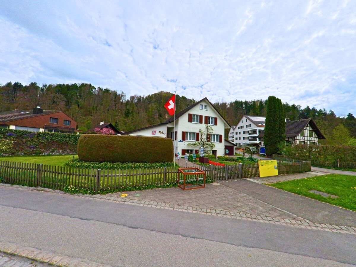 In Turbenthal (8488, Kanton Zürich) wirbt ein gelbes Schild vor einem gepflegten Einfamilienhaus mit Schweizer Fahne für einen bevorstehenden Flohmarkt, umgeben von weiteren Wohnhäusern und bewaldeten Hängen.