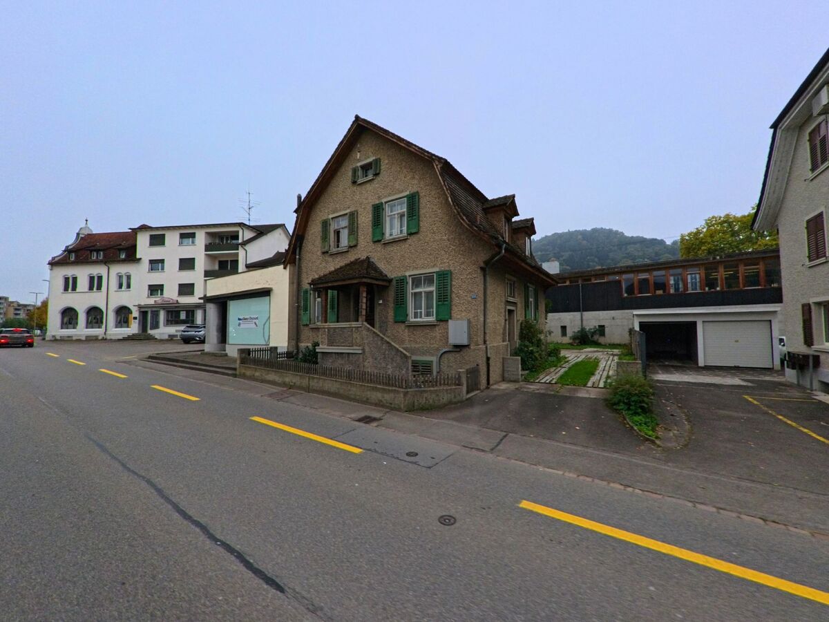 In Turgi (5300, Kanton Aargau) steht ein aelteres Wohnhaus mit gruennen Fensterlaeden direkt an der Strasse, flankiert von weiteren Gebaeuden und mit bewaldeten Huegeln im Hintergrund.